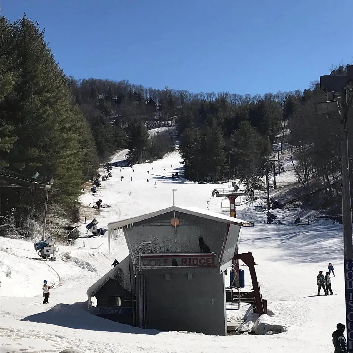 This image captures the base of the Wolf Ridge ski slopes, featuring a ski lift in motion, skiers descending the snowy runs, and the lodge building with a visible 'Wolf Ridge' sign. The snow-covered ground and bustling activity establish the sense of a true winter sports destination just steps away.