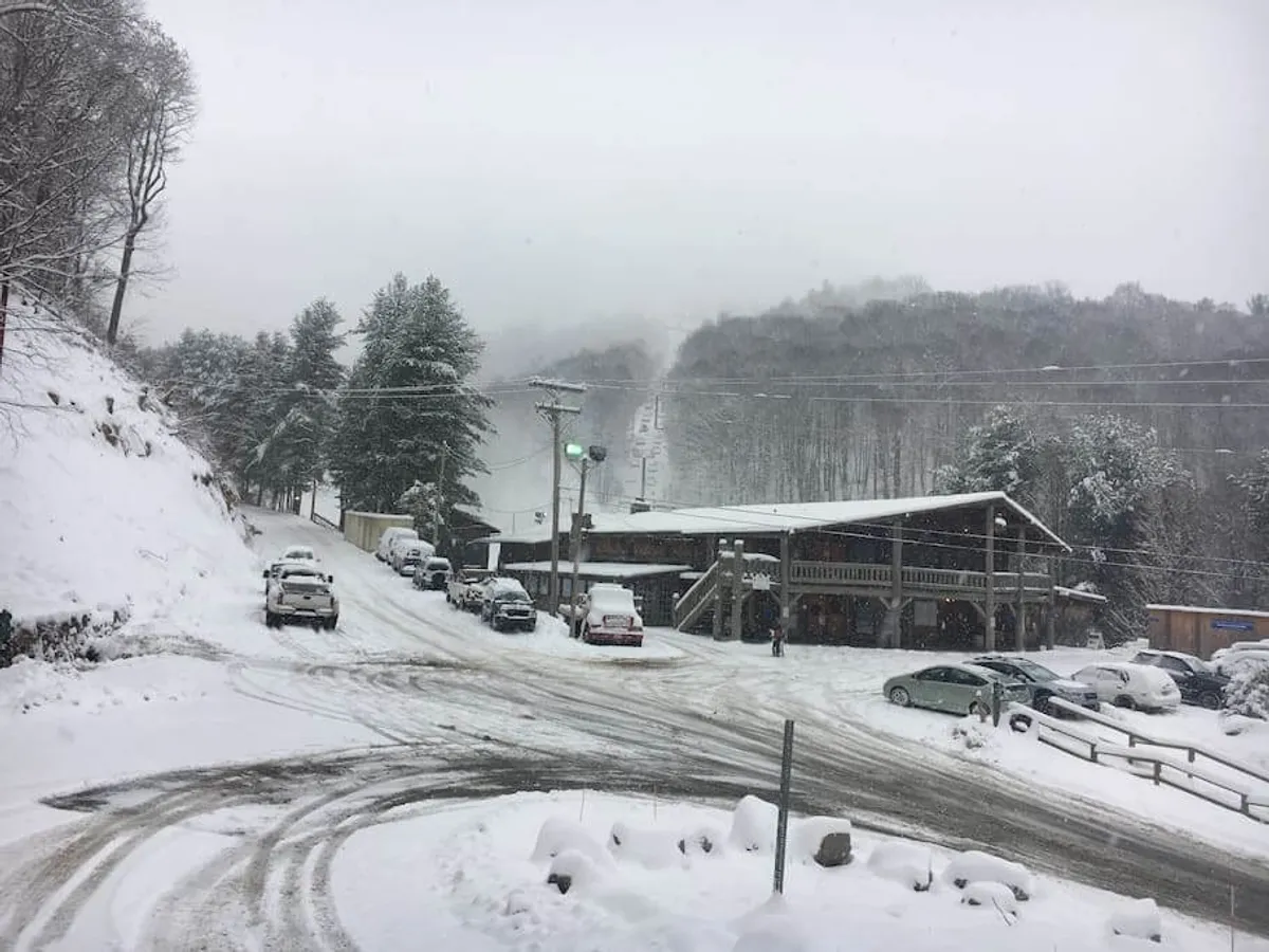 This exterior shot shows a snow-covered parking lot with cars, a wooden lodge structure, and surrounding trees with a mountain backdrop. The wintry scene reflects both accessibility and the cozy mountain ambiance.