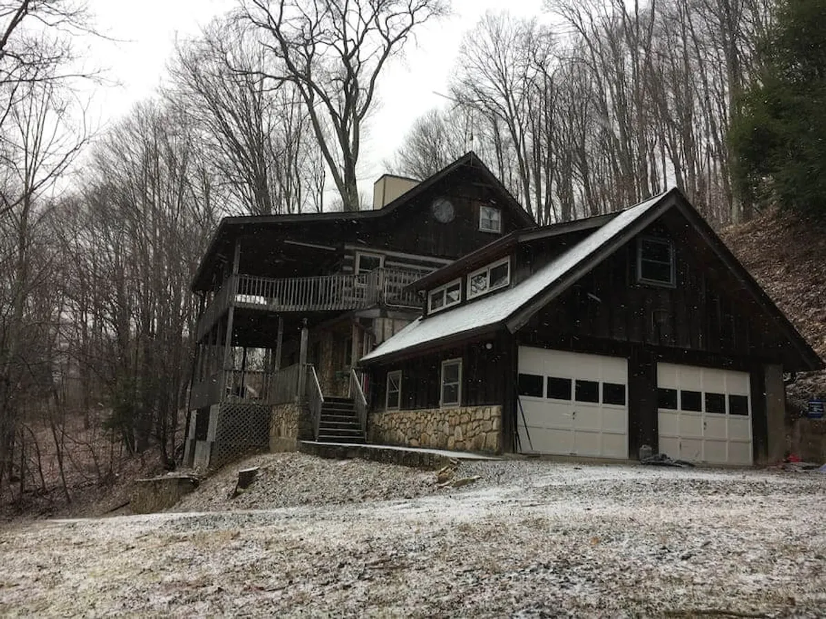 The image showcases a charming wooden cabin surrounded by snow, nestled amidst tall trees. The balcony faces a peaceful wintry scene, inviting guests to relax and soak in the mountain atmosphere. The home sits just steps from the ski lodge, making it the closest lodging option for winter adventure. The serene setting evokes warmth and togetherness, with the promise of cozy gatherings inside after a day in the snow.