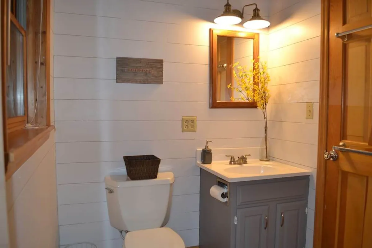 A bright bathroom scene with a well-lit mirror, a modern vanity, a decorative flower arrangement, and a wooden accent sign. The space feels fresh, organized, and tranquil, ideal for refreshing and recharging.