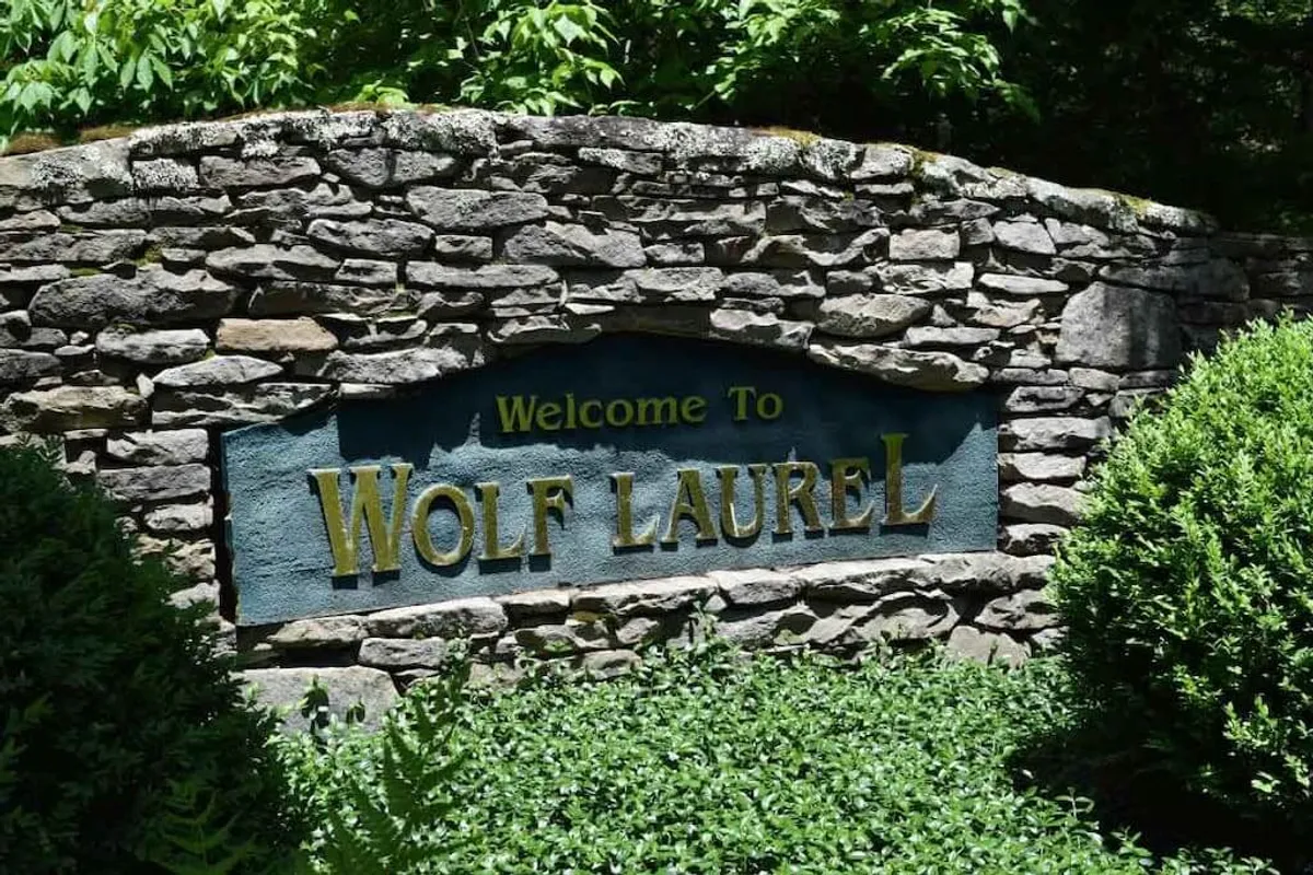 A stone entrance sign marks the entry to the Wolf Laurel community, surrounded by lush greenery and well-maintained landscaping. The image suggests a secure and welcoming entry point to the gated neighborhood.