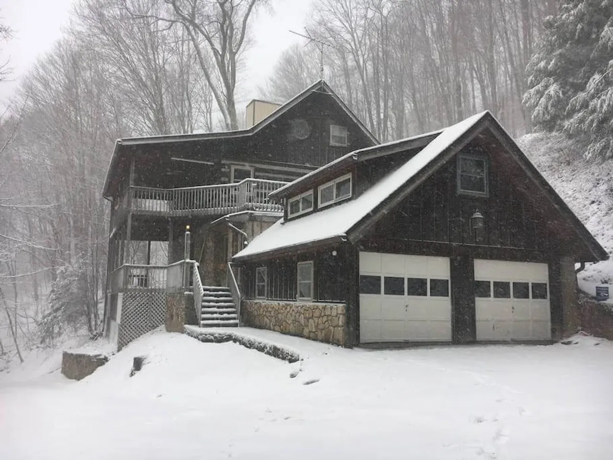 A serene winter exterior of the property, featuring a snow-covered roof, wooden balcony, sturdy stone foundation, and garage doors—set amidst a peaceful, private setting. The image conveys an inviting sense of seclusion within a secure, gated mountain community.
