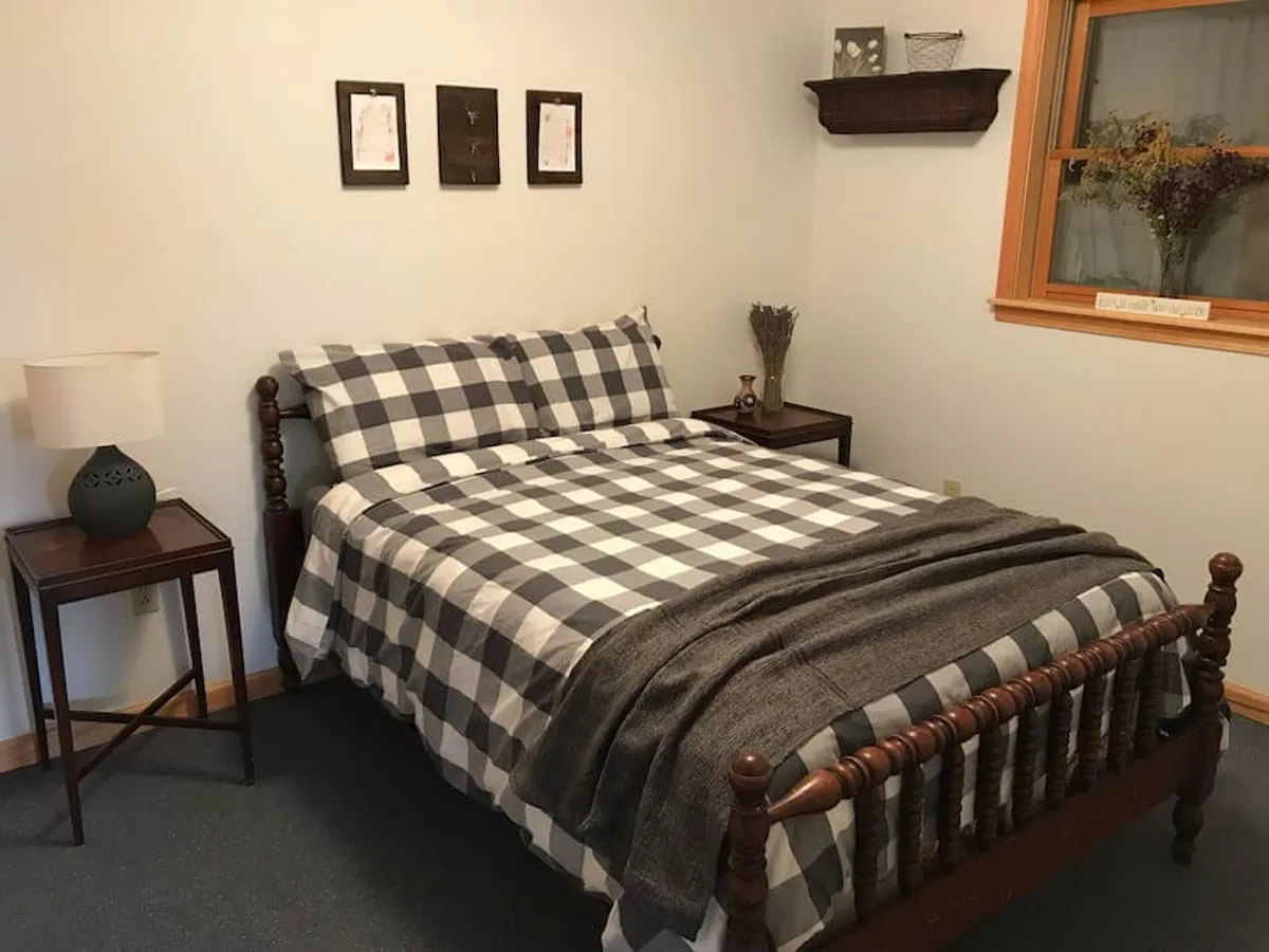 A cozy bedroom featuring a bed with checkered bedding, nightstands on either side, and framed artwork above the headboard. Soft light filters into the room, enhancing the peaceful and comfortable atmosphere.